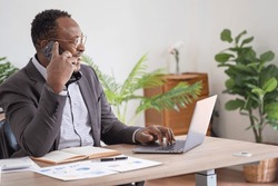 An African American businessman in a black suit uses a mobile phone to communicate with clients and a laptop to view company profits. to build confidence for customers to come and invest in the office