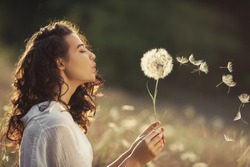 Beautiful Young Woman sitting on the field in green grass and blowing dandelion. Outdoors. Enjoy Nature. Healthy Smiling Girl on summer lawn. Allergy free concept. Gorgeous slim mixed race Caucasian