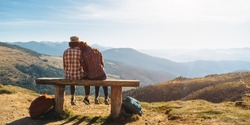Couple of hikers with backpacks enjoying valley landscape view from top of a mountain. Young adult tourists, man and woman sitting in the bench. Panoramic view of mountain hills, Carpathian mountains