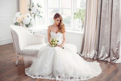 Wedding. Bride in beautiful dress sitting on sofa indoors in white studio interior like at home. Trendy wedding style shot in full length. Young attractive caucasian brunette model like a bride