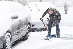 Young man, male in winter coat cleaning, shoveling driveway, street from covered falling, snow in heavy snowing snowstorm with shovel, residential houses, cars parked on road