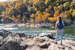 Young woman photographer with backpack and camera looking at view of Potomac river in Great Falls with autumn colorful foliage in Maryland