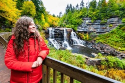 Young woman on wooden boardwalk deck overlook railing at Blackwater Falls waterfall state park in West Virginia autumn fall season looking at view happy