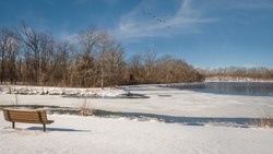 View of Midwestern lake partially covered with melting ice on nice spring day; wooden bench at the water edge; flock of  birds flying over lake
