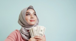 Happy Asian muslim woman with dollars in hand is thinking about the thing to buy. rich girl, finance and investment concept.