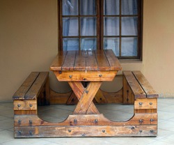 Sturdy wooden table with two seats attached as one unit standing on stoep against a window
