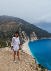 Smiling caucasian woman in long white beach shirt standing in the rain at bluff edge with Myrtos Beach in background. Popular travel destination in Cephalonia, Greece, Europe