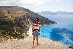 Smiling caucasian young woman in striped multicolored shirt and jeans shorts standing at bluff edge with Myrtos Beach in background. Popular travel destination in Cephalonia, Greece, Europe