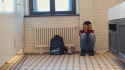 Depressed african child abandoned in lavatory and leaning against wall. Young afro boy sitting alone with sad feeling at school. Bullying, discrimination and racism concept