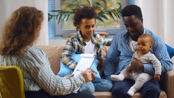 Social worker consulting smiling young single father with kids at home. Case worker woman visiting african man with schoolboy son and baby daughter providing consultation