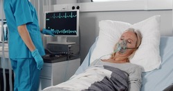 Doctor caring sick patient in hospital checking vital signs on monitor. Portrait of ill senior woman with oxygen mask resting in bed and nurse using computer on background checking physical condition