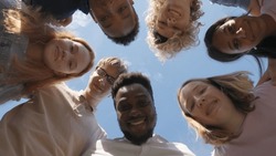 Low angle view of multiethnic teen students and teacher stand in circle and look down at camera outdoors. Professor with students hug and smile outside school