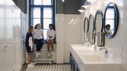 Multiethnic teen girlfriends chat in school bathroom during break. Diverse teenage students gossip in campus toilet after lesson