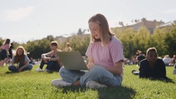 Schoolgirl using laptop while sitting on green grass of campus lawn. Pretty girl student preparing for classes in nature, her fellow students sitting in distance blurred on background. 