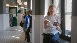 Teenage girl having lunch and surfing internet on smartphone sitting on windowsill at school during break. Teen student eat sandwich and chat on cellphone in campus