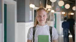 Pretty teen schoolgirl looking at camera and smiling standing in corridor. Happy teenage student with backpack and books posing at camera in campus