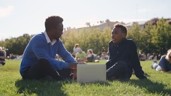 African-American male professor discussing exam results with student sitting on lawn near school. Portrait of teacher and teenage boy sitting on grass on schoolyard and talking with laptop
