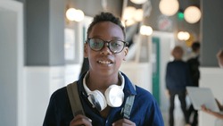 Portrait of teen African-American student in corridor wearing glasses, smiling at camera. Happy teenage student looking at camera standing in campus hallway