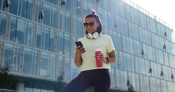 Low angle view fitness woman using phone with workout app exercising in city. Portrait of Indian female with bottle of water and smartphone after workout outdoors