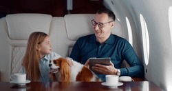 Mature businessman travelling on personal jet with little daughter and dog. Father using digital tablet and talking to preteen girl sitting in first class airplane interior