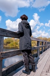 A girl in black clothes stands with her back on a wooden bridge on an autumn sunny day