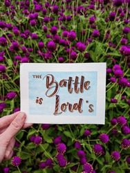 A hand holding a card written with hand-lettering style, saying The Battle is the Lord's with the pretty background of garden or flowers