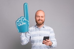 Charming handsome office man pointing with fan foam glove and holding smartphone over grey background