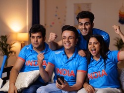 Thrilled friends in Indian jerseys cheering for team India during match. Excited cricket fans sitting at home watching game on TV, using smartphone app to online bet, friends celebrating victory