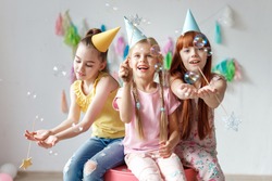 Portrait of three beautiful girls wear festive caps, play with bubbles, sit together on chair, celebrate birthday, being in good mood, use magic wand, have party in decorated room. Childhood concept