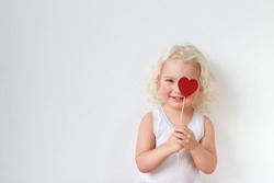 Blue eyed pretty small kid dressed casually, has fun indoors, covers eye with heart stick, glad to spend free time with her parents, isolated over white background with copy space for your text