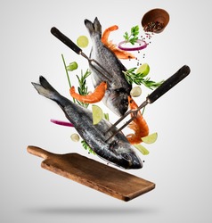 Flying raw whole bream fish and prawns, with ingredients for cooking. Freeze motion. Fork holding the meat. Concept of food preparation in low gravity mode. Separated on smooth gray background