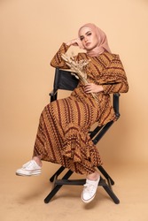 Beautiful female model wearing modern batik kebaya with hijab, sitting on a director chair isolated over beige background. Stylish Muslim female fashion lifestyle portraiture concept.