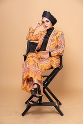 Beautiful female model wearing modern batik kebaya with turban style hijab, sitting on a director chair isolated over beige background. Stylish Muslim female fashion lifestyle portraiture concept.
