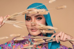Close up model model wearing modern batik kebaya with hijab, holding a dried flower isolated over beige background. Stylish Muslim female fashion lifestyle  concept.