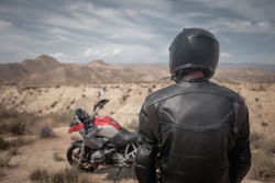 A man in black helmet and black leather jacket standing near adventure motorcycle in desert, adventure overland travel, back view