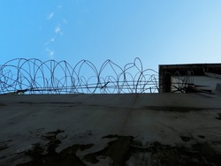 High concrete fence with peeling plaster and barbed wire against the blue sky. Bottom view