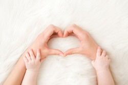 Heart shape created from young woman fingers. Infant hands on mother arms. White, soft, fluffy fur carpet background. Lovely emotional, sentimental moment. Closeup. Point of view shot.