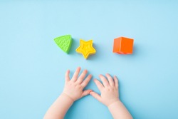Baby hands playing with green triangle, yellow star and orange square shapes on light blue table background. Pastel color. Closeup. Toys of development for little kids.