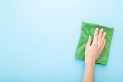 Young adult woman hand holding green rag and wiping table, wall or floor surface in kitchen, bathroom or other room. Closeup. Empty place for text or logo. Light blue background. Pastel color.