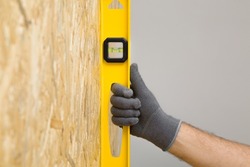 Young adult man hands using yellow spirit level and measuring vertical surface of osb board for new wall. Closeup. Preparing for repair work of home. Renovation process.