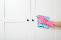 Young adult woman hand in pink rubber protective glove using blue dry rag and wiping white wooden doors of wardrobe. Closeup. Regular cleanup at home. Front view.