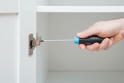 Young adult man hand using manual screwdriver and screwing screw in hinge for white wardrobe door. Assembling new wooden furniture. Closeup.
