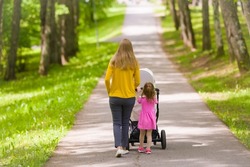 Young adult mother and little daughter pushing white baby stroller and walking at town park in warm sunny summer day. Spending time together and breathing fresh air. Enjoying stroll. Two child mom.