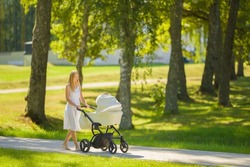 Smiling young adult mother in dress pushing white baby stroller and walking on sidewalk at town park in warm sunny summer day. Spending time with newborn and breathing fresh air. Enjoying stroll.