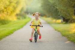 Happy beautiful little girl fast running and riding on first bike without pedals on sidewalk at city park in warm summer day. Cute 3 years old toddler. Front view. Learning to keep balance.