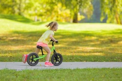 Happy beautiful little girl fast running and riding on first bike without pedals on sidewalk at city park in warm summer day. Cute 3 years old toddler. Side view. Learning to keep balance.