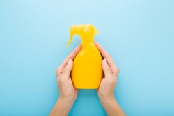 Young adult woman hands holding yellow spray bottle of sunscreen or cleaning detergent on light blue table background. Pastel color. Closeup. Point of view shot.