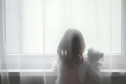Little girl holding white teddy bear standing alone at window behind transparent day curtains and looking out from home. Back view. Waiting concept. Close up.
