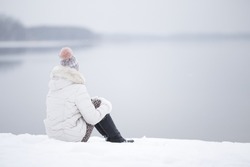 Young adult woman sitting alone on snow at lake shore and looking far away. White cold snowy winter day. Thinking about life. Peaceful atmosphere in nature. Back view.