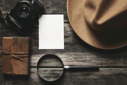 Mysterious detective game concept notebook leather cover sheet white paper felt brown hat portable mirrorless microthird digital photo camera big vintage magnifier isolated on black aged wood table